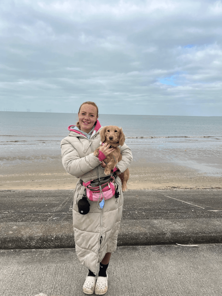 Amy standing on a beach promenade holding a dachshund